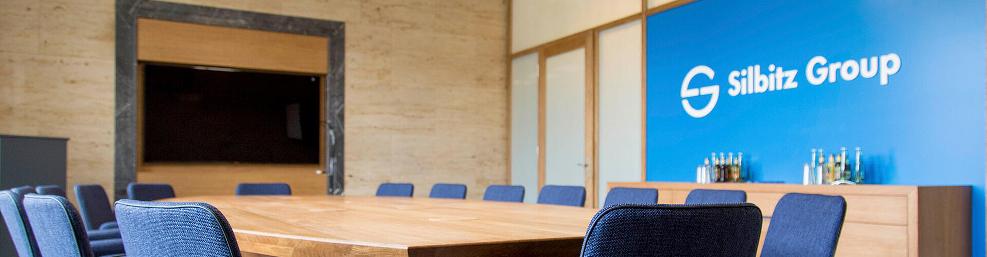 Ein Meeting Room mit großem, rundem Tisch aus Holz. Dahinter an einer blauen Wand ist das Silbitz Group Logo zu sehen.
