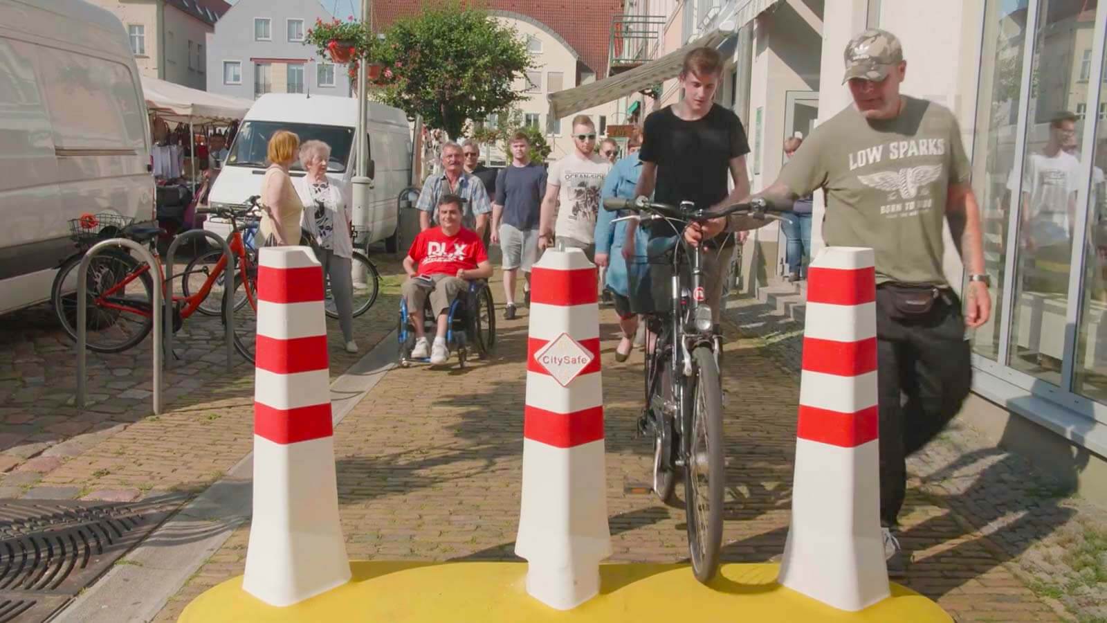 Ein CitySafe Anti-Terror-Poller steht in einer Fußgängerzone. Eine Gruppe Fahrradfahrer schieben gerade Ihre Fahrräder durch die Sperre.