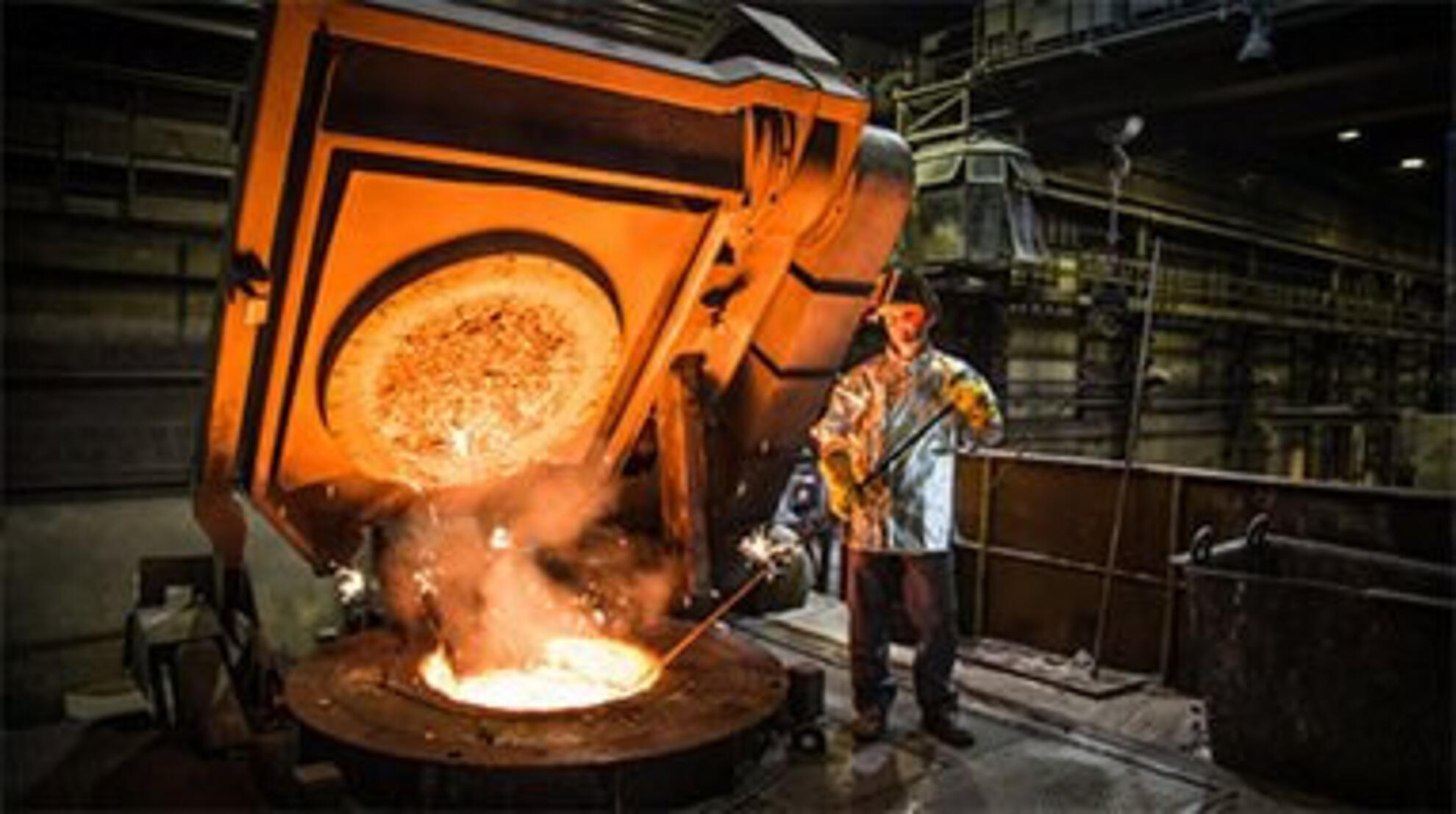 Arbeiter in Schutzkleidung überwacht in einer Gießerei, wie ein großer Schmelzofen glühend flüssiges Gusseisen in eine runde Form gegossen wird.