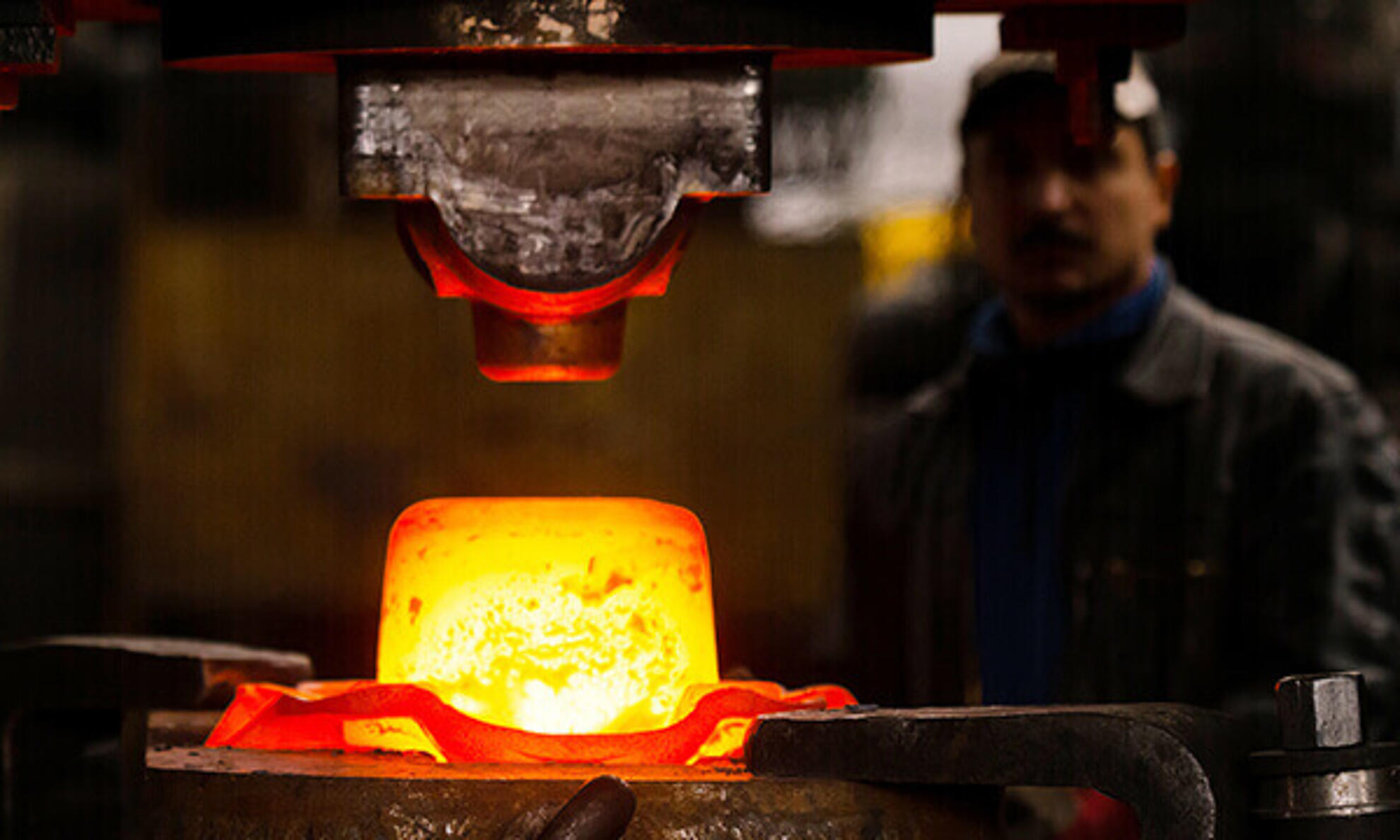 Ein glühendes Metallstück liegt unter einem hydraulischen Hammer, der von einem Mitarbeiter gesteuert wird.